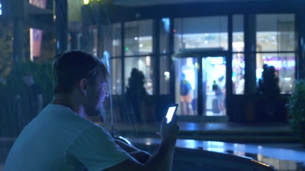 Молодий красивий хлопець сидить ввечері біля фонтану з кольоровим водяним освітленням. розмовляючи по телефону, розмивання, 4k — стокове відео