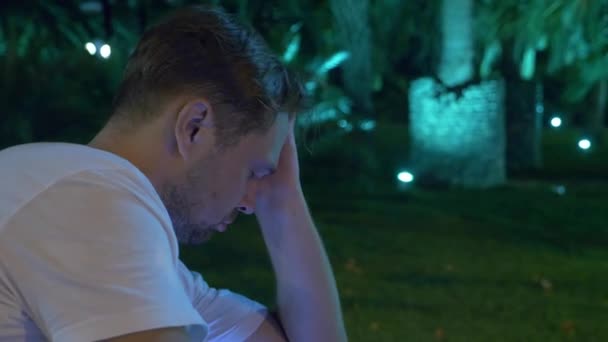 Homem sentado à noite no jardim tropical, debaixo de uma palmeira. 4K. escuro — Vídeo de Stock