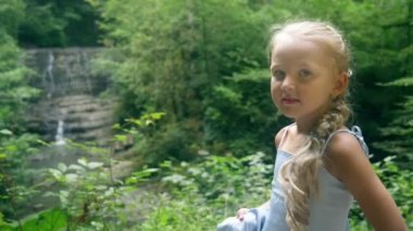 bir şelale orman karşı poz güzel mavi elbiseli küçük kız modeli. 4k, ağır çekim.