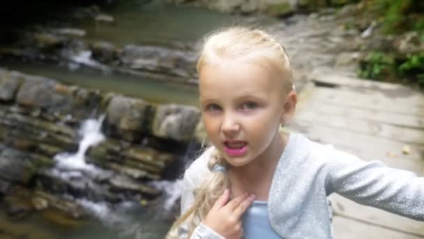 Primer plano. retrato de una niña modelo en un hermoso vestido azul, posando contra una cascada en un bosque. hace caras graciosas. 4k, cámara lenta . — Vídeo de stock