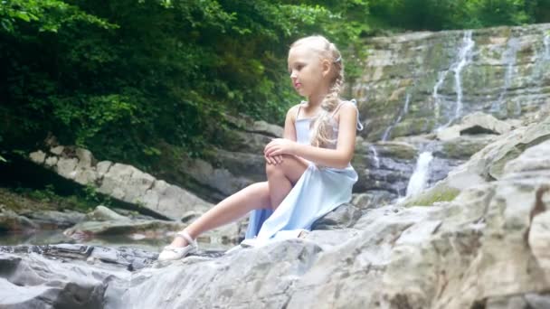 Modello bambina in un bellissimo vestito blu, seduto contro una cascata in una foresta. . 4k, rallentatore . — Video Stock