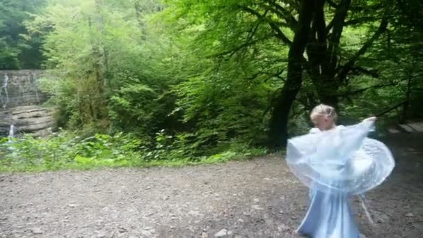 Petite fille modèle dans une belle robe bleue posant contre une cascade dans la forêt. 4k, au ralenti . — Video