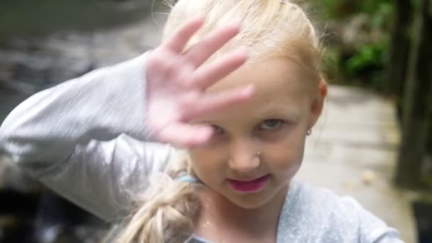 Крупным планом. портрет маленькой девочки-модели в красивом голубом платье, позирующей против водопада в лесу. строит смешные рожи. 4k, slow motion . — стоковое видео