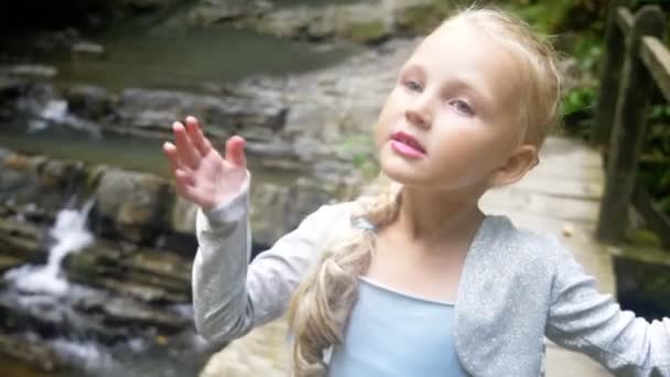 Nahaufnahme. Porträt eines kleinen Mädchens Modell in einem schönen blauen Kleid, posiert vor einem Wasserfall in einem Wald. macht lustige Gesichter. 4k, Zeitlupe. — Stockvideo