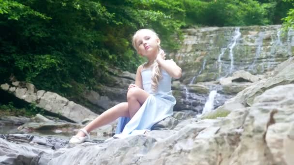 Маленькая девочка-модель в красивом голубом платье, сидящая против водопада в лесу. - Да. 4k, slow motion . — стоковое видео