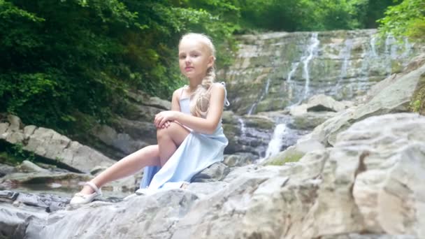 Meisje model in een mooie blauwe jurk, zittend tegen een waterval in een forest. . 4k, slow-motion. — Stockvideo