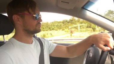 Güneş gözlüklü genç yakışıklı bir adam bir araba dağ manzaralı bir otoyol boyunca yüksek hızda yol açar. 4k, ağır çekim