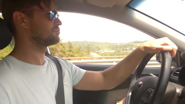 Молодой красивый мужчина в солнечных очках ведет машину на высокой скорости, вдоль шоссе с видом на горы. 4k, slow motion — стоковое видео