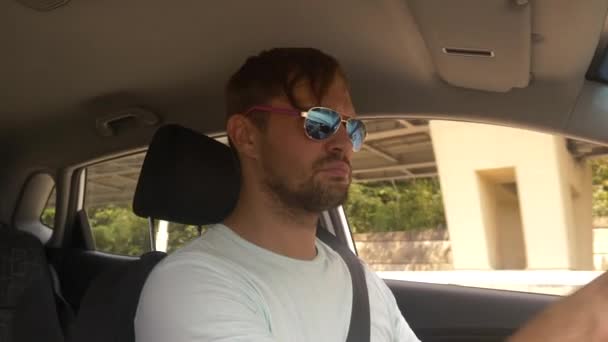 Молодий красивий чоловік в сонцезахисних окулярах веде машину на високій швидкості, вздовж шосе з видом на гори. 4k, повільний рух — стокове відео