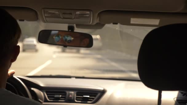 Ένας όμορφος νεαρός με γυαλιά ηλίου οδήγηση ενός αυτοκινήτου γύρω από την πόλη. αντανάκλαση σε το κάτοπτρο οδηγήσεως. 4k, αργή κίνηση — Αρχείο Βίντεο