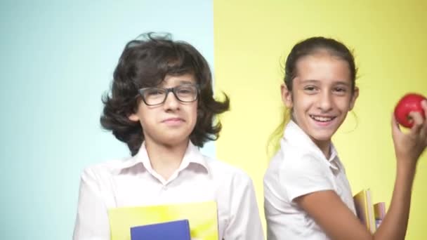 Çocuklar okulda bir renkli arka plan üzerinde üniforma portreleri. Komik çocuk. Kız ve erkek kardeş. öğrenme kavramı. Onlar kitap ve elma ellerinde, bak kamera ve gülümseme idare. — Stok video