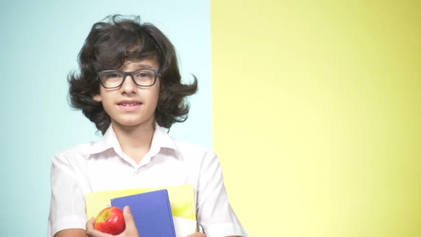 Retratos de um adolescente de uniforme escolar sobre fundo colorido. Um tipo engraçado. conceito de aprendizagem. Um adolescente está segurando livros olhando para a câmera e sorrindo. espaço de cópia — Vídeo de Stock