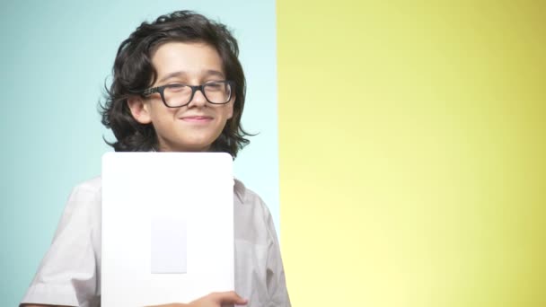 Porträtt av en tonåring i skoluniform och glasögon på en färgad bakgrund. Rolig kille. begreppet lärande. En tonåring håller en bärbar dator, tittar på kameran, leende och göra roliga ansikten — Stockvideo