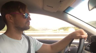Güneş gözlüklü genç yakışıklı bir adam bir araba dağ manzaralı bir otoyol boyunca yüksek hızda yol açar. 4k, ağır çekim