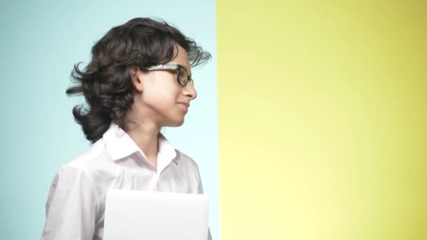 Portraits of a teenager in school uniform and glasses on a colored background. Funny guy. concept of learning. A teenager is holding a laptop, looking at the camera, smiling and making funny faces — Stock Video