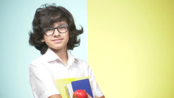 Retratos de um adolescente de uniforme escolar sobre fundo colorido. Um tipo engraçado. conceito de aprendizagem. Um adolescente está segurando livros olhando para a câmera e sorrindo. espaço de cópia — Vídeo de Stock