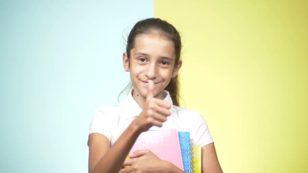 Portretten van een tienermeisje in school uniform op een gekleurde achtergrond. Grappig meisje. concept van leren. Een tiener houdt boeken kijken naar de camera en glimlachen. kopie ruimte — Stockvideo