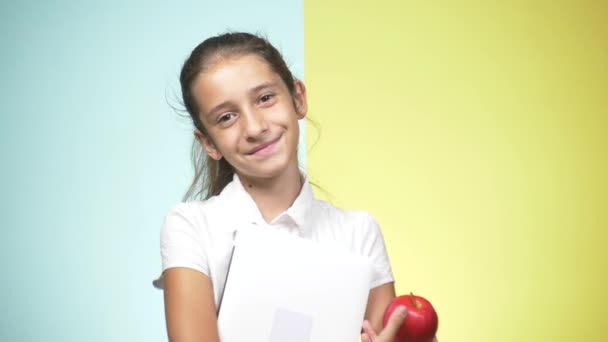 Retratos de uma adolescente de uniforme escolar sobre fundo colorido. Miúda engraçada. conceito de aprendizagem. Um adolescente está segurando livros olhando para a câmera e sorrindo. espaço de cópia — Vídeo de Stock