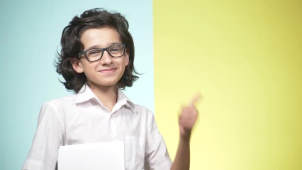 Retratos de um adolescente em uniforme escolar e óculos em um fundo colorido. Um tipo engraçado. conceito de aprendizagem. Um adolescente está segurando um laptop, olhando para a câmera, sorrindo e fazendo caras engraçadas — Vídeo de Stock