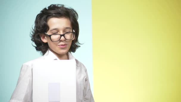 Retratos de um adolescente em uniforme escolar e óculos em um fundo colorido. Um tipo engraçado. conceito de aprendizagem. Um adolescente está segurando um laptop, olhando para a câmera, sorrindo e fazendo caras engraçadas — Vídeo de Stock