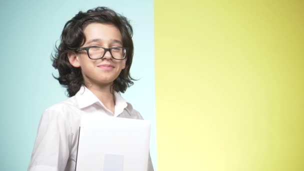 穿着校服的少年肖像和彩色背景的眼镜。有趣的家伙。学习的概念。一个十几岁的孩子拿着一台笔记本电脑, 看着相机, 微笑着, 做着滑稽的面孔。 — 图库视频影像