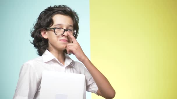 Retratos de un adolescente en uniforme escolar y gafas sobre un fondo de color. Qué gracioso. concepto de aprendizaje. Un adolescente sostiene un portátil, mira a la cámara, sonríe y hace caras graciosas — Vídeo de stock