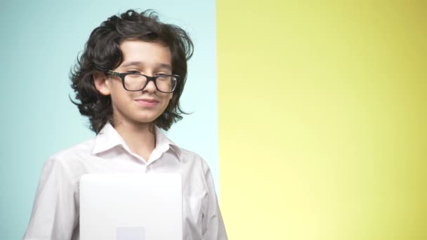 Retratos de un adolescente en uniforme escolar y gafas sobre un fondo de color. Qué gracioso. concepto de aprendizaje. Un adolescente sostiene un portátil, mira a la cámara, sonríe y hace caras graciosas — Vídeo de stock