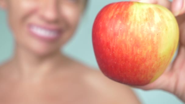 Primer plano, una chica con una sonrisa de diente blanco sobre un fondo azul sostiene una manzana roja. Concéntrate en la manzana, borrosa. 4k, cámara lenta — Vídeos de Stock