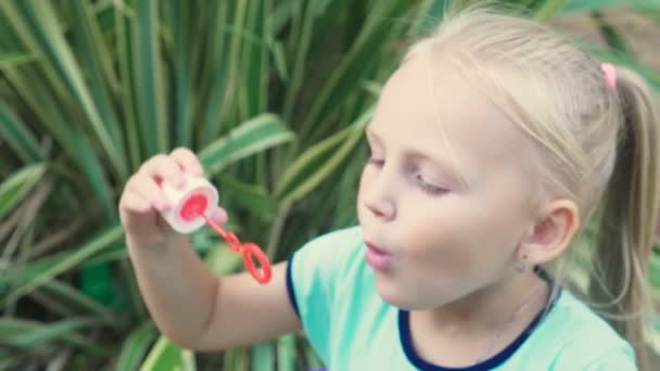 Little blond girl blowing soap bubbles against a tropical park background. 4k, slow motion — Stock Video