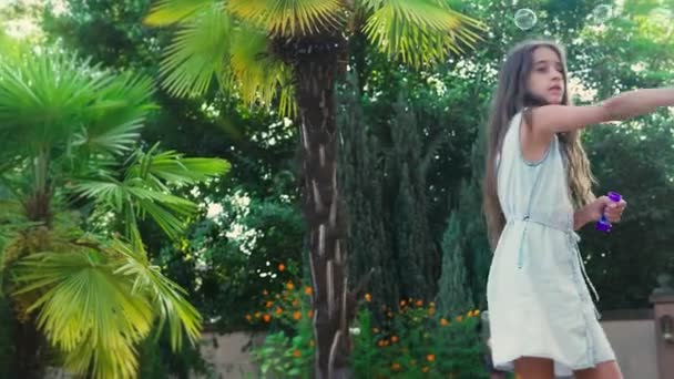 Брюнетка-подросток, пускающая мыльные пузыри на фоне тропического парка. 4k, slow motion — стоковое видео