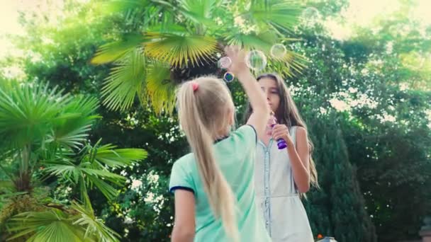 Due ragazze, bruna e bionda che soffiano bolle di sapone su uno sfondo di parco tropicale. 4k, rallentatore — Video Stock