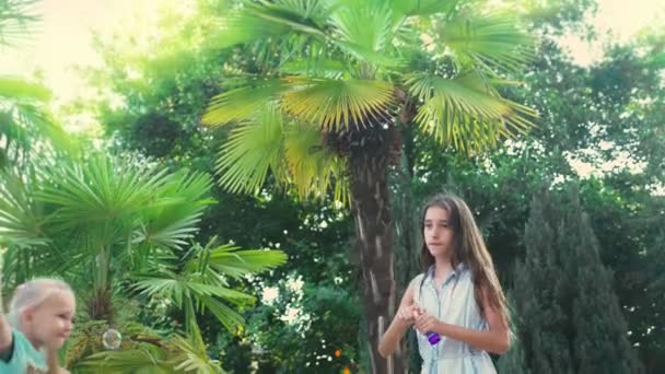Dwie dziewczyny, brunetka i blond, dmuchanie baniek mydlanych na tle tropikalnego parku. 4k, zwolnionym tempie — Wideo stockowe