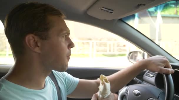 El hombre al volante del coche, come comida rápida, mientras el coche se mueve. en un día soleado de verano. 4k, cámara lenta — Vídeo de stock