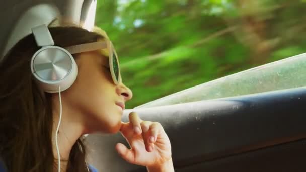 Malá elegantní dívka v klobouku a sluneční brýle poslechu hudby ve sluchátkách od jejího smartphone, ona jezdí v autě na zadním sedadle. 4k, pomalý pohyb
