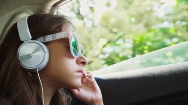 Una pequeña chica con estilo en un sombrero y gafas de sol escuchando música en los auriculares desde su teléfono inteligente, que monta en el coche en el asiento trasero. 4k, cámara lenta — Vídeo de stock