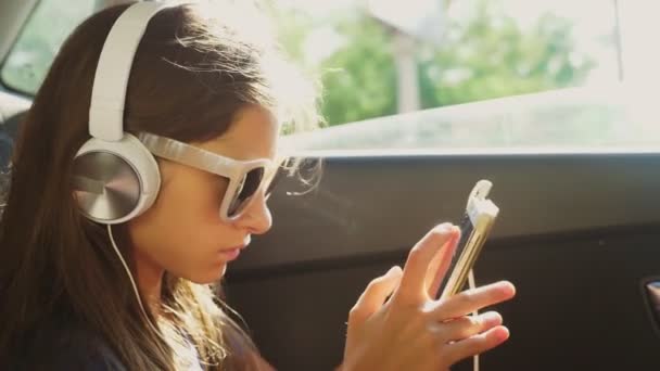 Una pequeña chica con estilo en un sombrero y gafas de sol escuchando música en los auriculares desde su teléfono inteligente, que monta en el coche en el asiento trasero. 4k, cámara lenta — Vídeos de Stock
