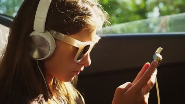 Una pequeña chica con estilo en un sombrero y gafas de sol escuchando música en los auriculares desde su teléfono inteligente, que monta en el coche en el asiento trasero. 4k, cámara lenta — Vídeos de Stock