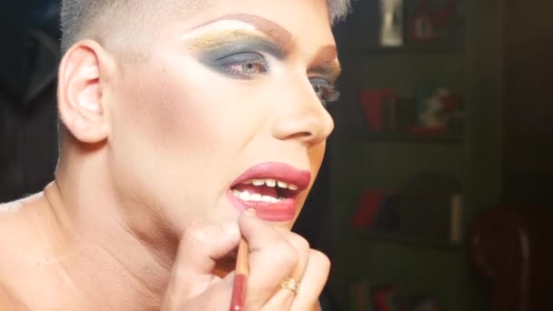 Begreppet parodi make-up. en stilig ung man applicerar makeup i ansiktet, sitter framför en spegel i omklädningsrummet. professionell travesti konstnär. närbild, 4k. — Stockvideo