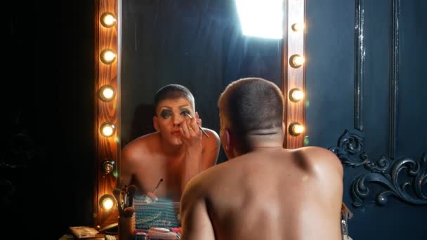 Konzept des Travestie-Make-ups. Ein gutaussehender junger Mann schminkt sein Gesicht und sitzt in der Garderobe vor einem Spiegel. professioneller Travestiekünstler. Nahaufnahme, 4k. — Stockvideo