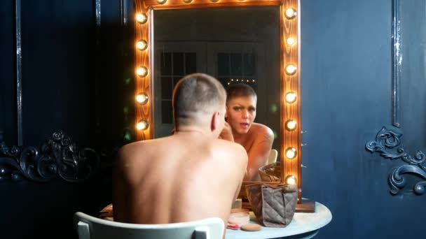 Travesti makyaj kavramı. yakışıklı bir genç adam soyunma odasında bir ayna önünde oturan yüzünde makyaj uygulanır. profesyonel karikatür sanatçısı. yakın çekim, 4k. — Stok video