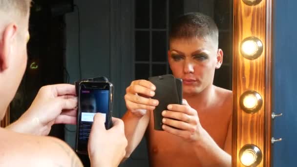 Begreppet parodi make-up. en stilig ung man applicerar makeup i ansiktet, sitter framför en spegel i omklädningsrummet. professionell travesti konstnär. närbild, 4k. — Stockvideo