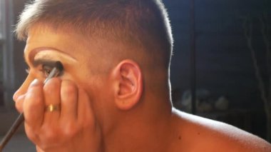 travesti makyaj kavramı. yakışıklı bir genç adam soyunma odasında bir ayna önünde oturan yüzünde makyaj uygulanır. profesyonel karikatür sanatçısı. yakın çekim, 4k.