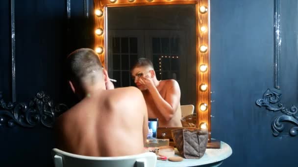 Έννοια του make-up παρωδία. ένας όμορφος νεαρός άνδρας ισχύει μακιγιάζ στο πρόσωπό του, κάθεται μπροστά από έναν καθρέφτη στο καμαρίνι. παρωδία επαγγελματίας καλλιτέχνης. γκρο πλαν, 4k. — Αρχείο Βίντεο