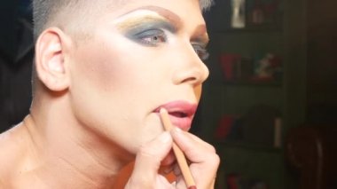 travesti makyaj kavramı. yakışıklı bir genç adam soyunma odasında bir ayna önünde oturan yüzünde makyaj uygulanır. profesyonel karikatür sanatçısı. yakın çekim, 4k.