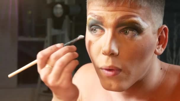 Concept de travestissement maquillage. un beau jeune homme applique du maquillage sur son visage, assis devant un miroir dans le dressing. artiste de parodie professionnelle. gros plan, 4k . — Video