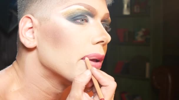 Begreppet parodi make-up. en stilig ung man applicerar makeup i ansiktet, sitter framför en spegel i omklädningsrummet. professionell travesti konstnär. närbild, 4k. — Stockvideo