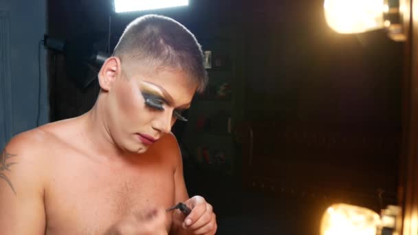 Conceito de maquiagem travesti. um jovem bonito aplica maquiagem em seu rosto, sentado na frente de um espelho no camarim. artista travesti profissional. close-up, 4K . — Vídeo de Stock