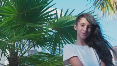 güzel esmer kız palm bir arka plan karşı poz bırakır. Mavi uzun kıvırcık saçlı bir kız Genç. yaz güneşli gün. 4k, ağır çekim