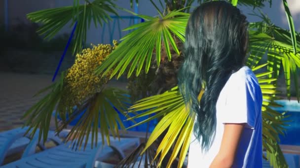 Piękna brunetka dziewczyna pozowanie na tle palm liści. Nastolatek dziewczyna z niebieski długie kręcone włosy. słoneczny letni dzień. 4k, zwolnionym tempie — Wideo stockowe