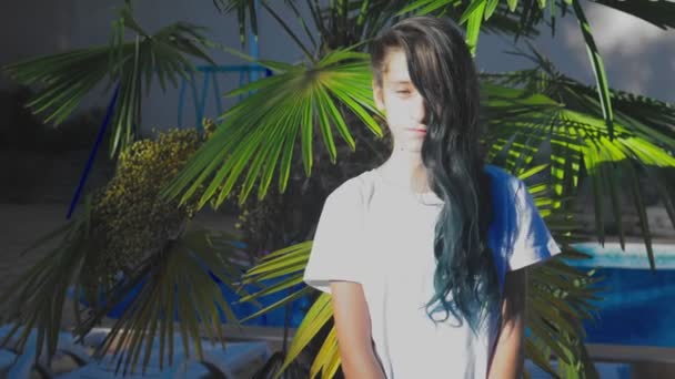 Güzel esmer kız palm bir arka plan karşı poz bırakır. Mavi uzun kıvırcık saçlı bir kız Genç. yaz güneşli gün. 4k, ağır çekim — Stok video